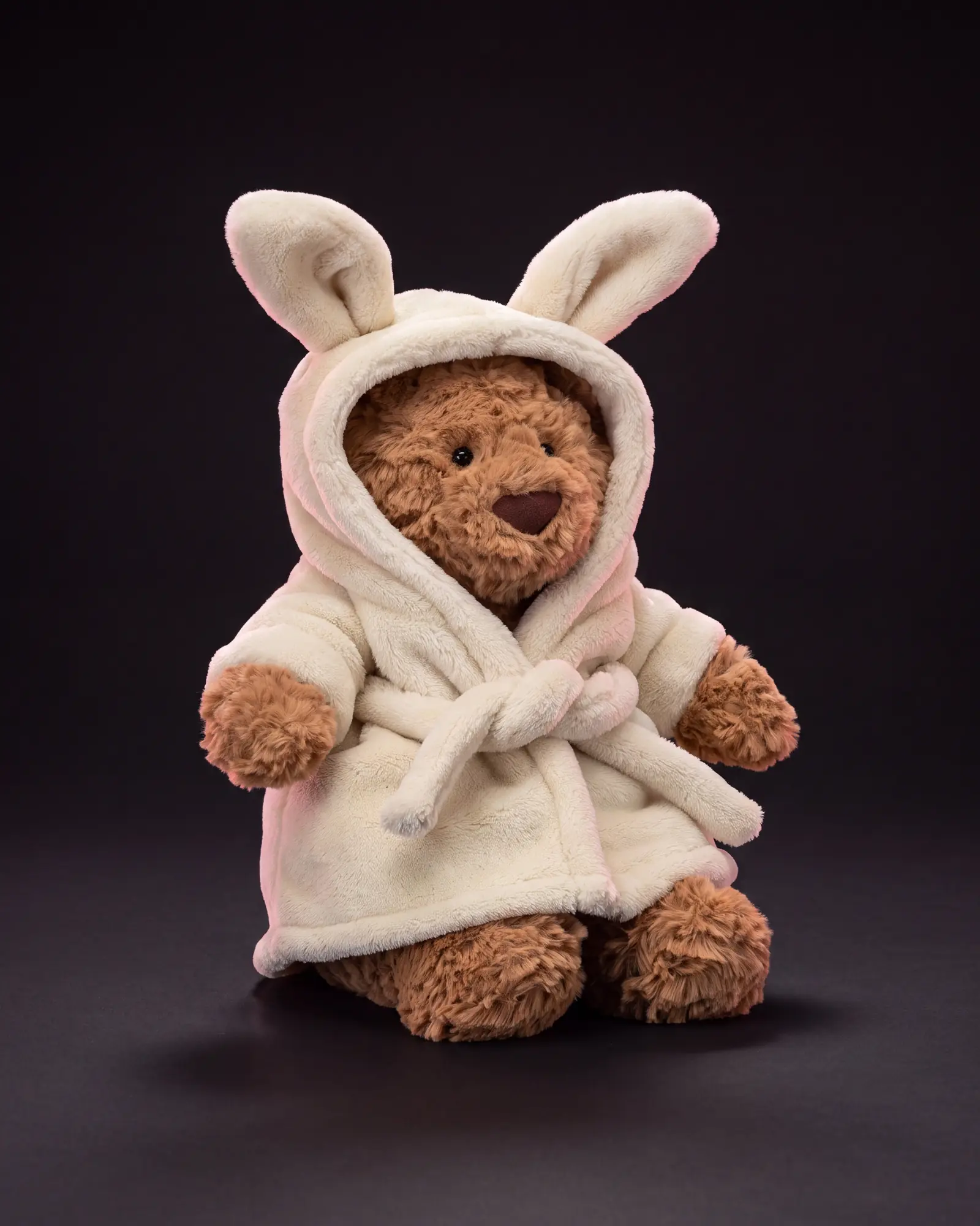 A golden-brown Jellycat bear in a cream hooded bathrobe with rabbit ears, on a dark studio sweep.