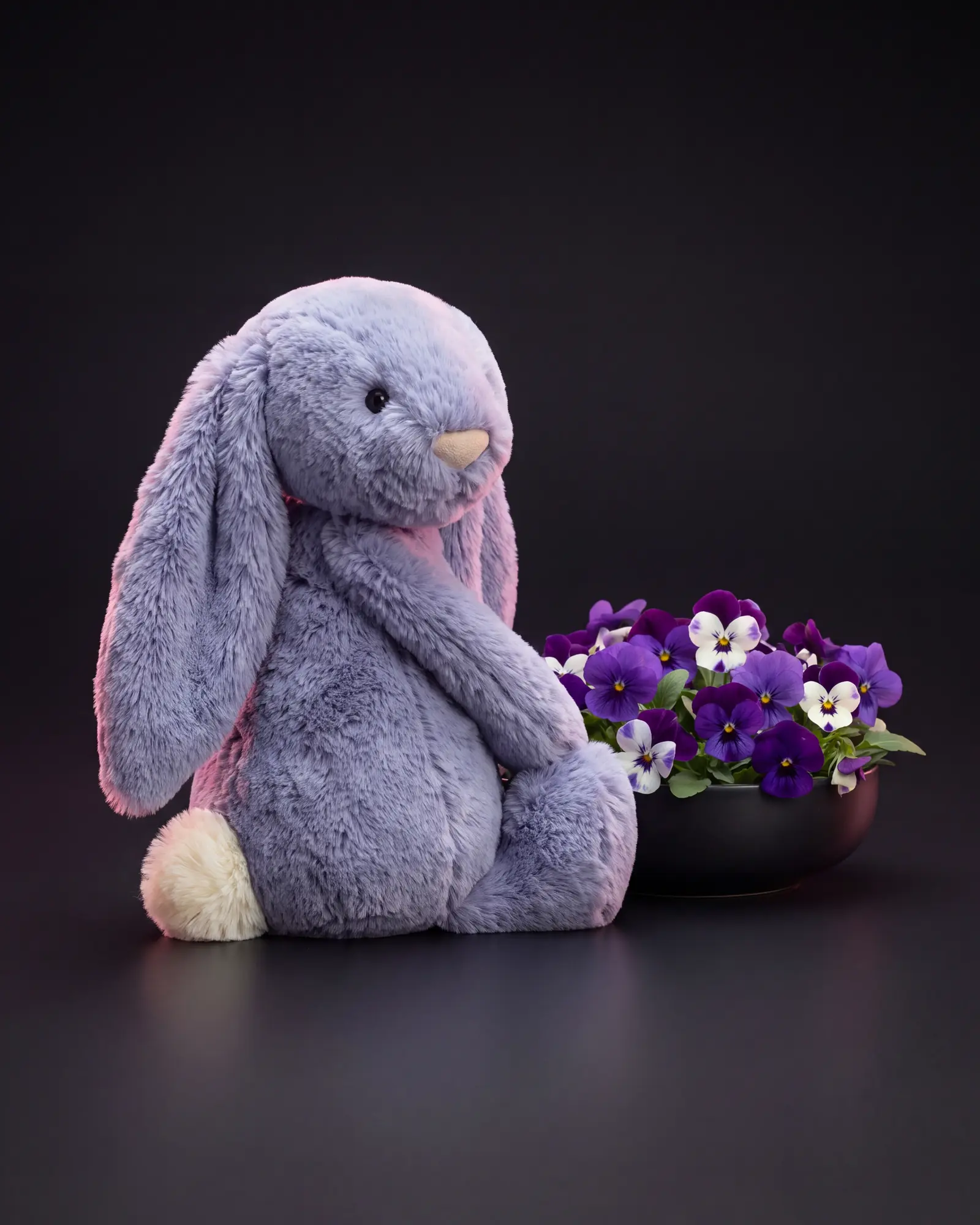 The bunny looking back over its shoulder beside a low dark dish of violet and white viola flowers.