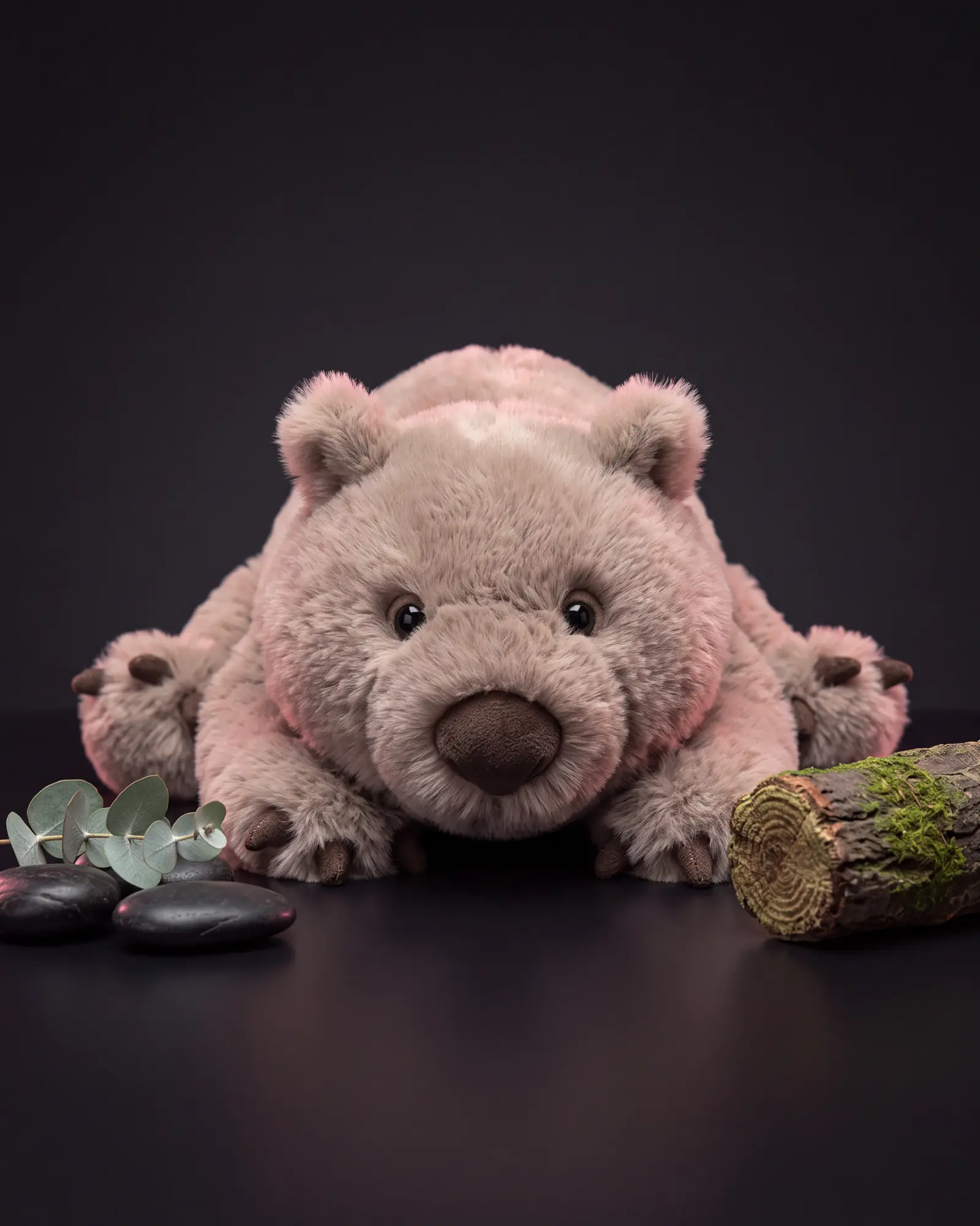 The wombat lying flat on its belly with its legs sprawled out, beside a mossy log, dark river stones and a sprig of eucalyptus.